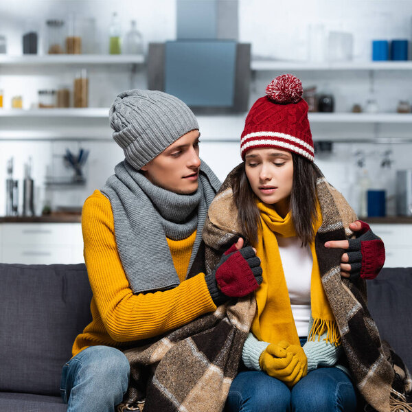 young man in knitted hat and gloves covering cold girlfriend with warm plaid blanket in kitchen