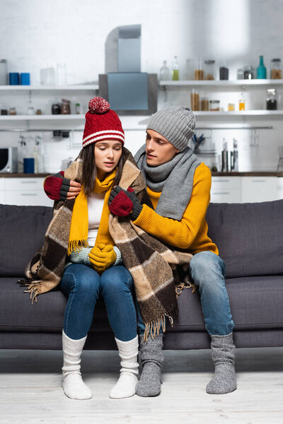 young man in warm hat and gloves covering freezing girlfriend with plaid blanket in cold kitchen
