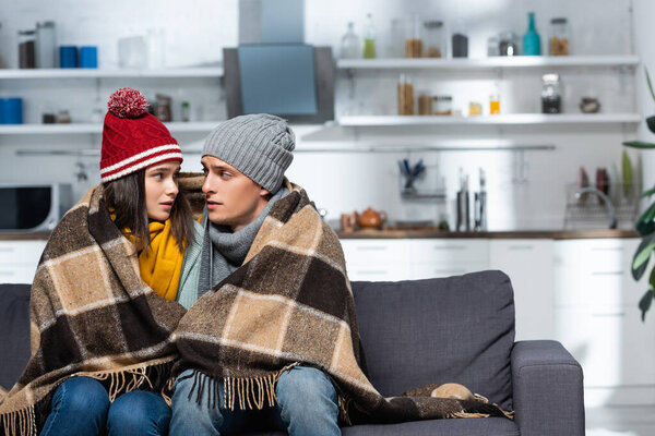 freezing couple in warm hats looking at each other while sitting under plaid blanket in cold kitchen