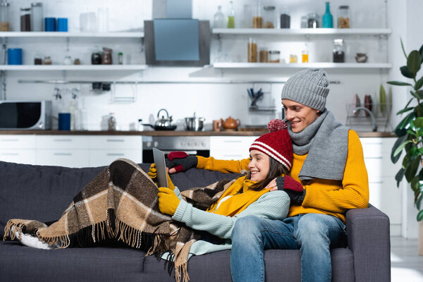 excited couple in warm hats and gloves watching movie on laptop in cold kitchen
