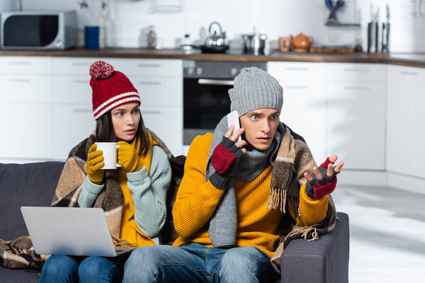 беспокойный мужчина разговаривает на смартфоне рядом с холодной женщиной в теплых перчатках шляпа над головой, сидя с ноутбуком в холодной кухне