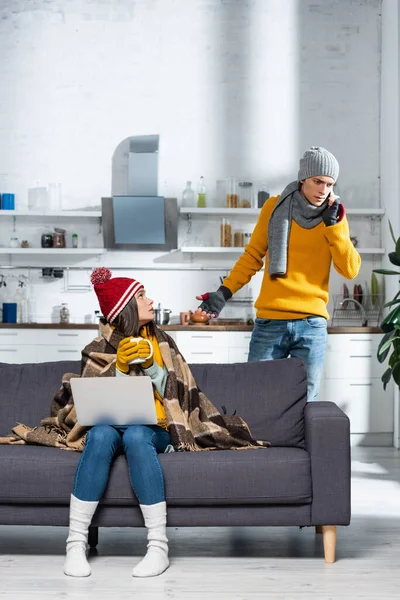 暖かい帽子と手袋の凍った女性がノートパソコンでソファに座っている間に緊張したボーイフレンドがスマートフォンで話している間 — ストック写真