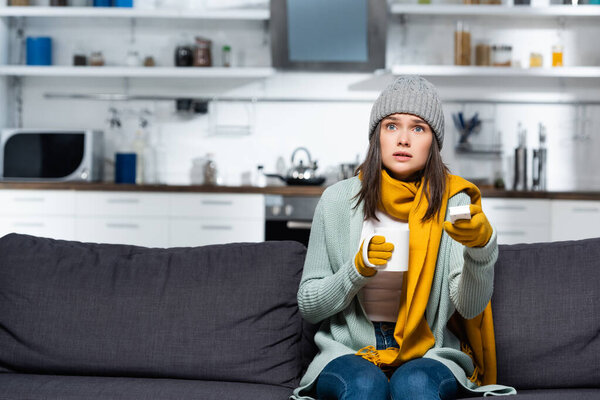 frozen woman in knitted clothes holding tv remote controller and cup of warming beverage while sitting on sofa in cold kitchen
