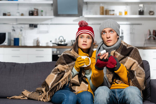 freezing couple in warm hats, wrapped in plaid blanket, watching tv while sitting in cold kitchen