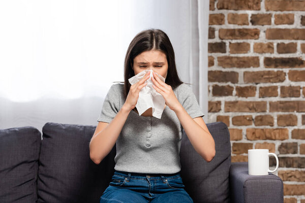 diseased woman sneezing in paper napkin while sitting on sofa near cup of warming beverage