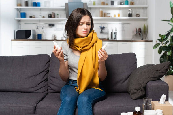 diseased woman with scarf on neck holding nasal and throat sprays while sitting on sofa in kitchen