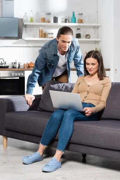 Man Standing Joyful Girlfriend Sitting Sofa Using Laptop Kitchen — Stock Photo, Image
