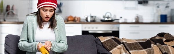 website header of freezing woman in knitted hat putting on warm gloves while sitting on sofa in cold kitchen