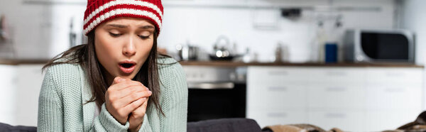 horizontal image of freezing woman in knitted hat blowing on clenched hands in cold kitchen