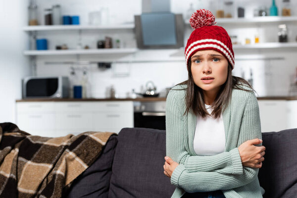 freezing woman in knitted hat hugging herself while sitting in cold kitchen