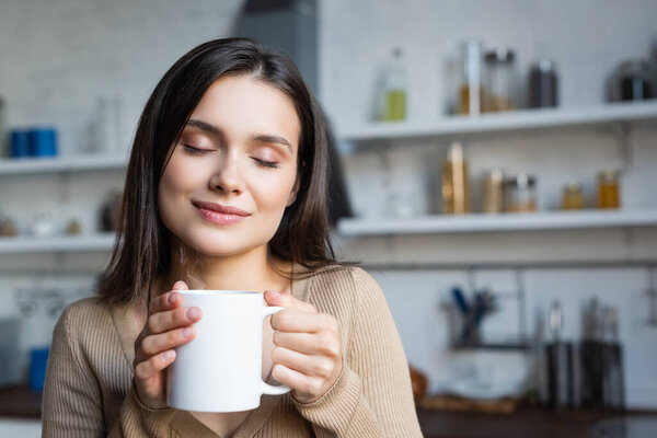 pleased young woman with closed eyes holding cup of warm tea at home