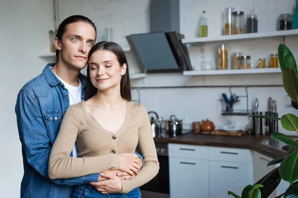 young man hugging woman standing in kitchen with closed eyes