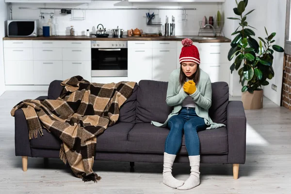 Shivering woman in warm hat, gloves and socks sitting on sofa near plaid blanket in kitchen — Stock Image Shivering Woman Warm Hat Gloves Socks Sitting Sofa Plaid Blanket Stock Picture