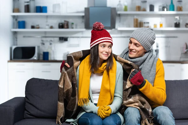 Young man in warm hat and gloves covering freezing girlfriend with plaid blanket while sitting on sofa in cold kitchen — Stock Image Young Man Warm Hat Gloves Covering Freezing Girlfriend Plaid Blanket Royalty Free Stock Images