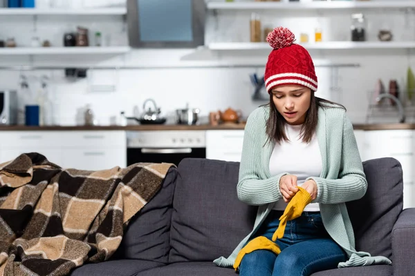Cold woman in knitted hat putting on warm gloves while sitting on couch in kitchen — Stock Image Cold Woman Knitted Hat Putting Warm Gloves While Sitting Couch Stock Photo