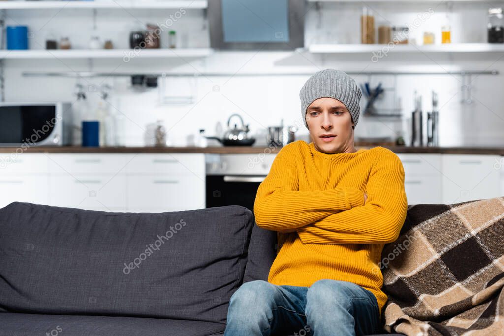 shivering man in knitted sweater and hat sitting on sofa with crossed arms in cold kitchen