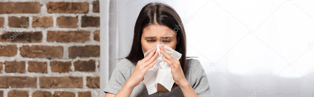 horizontal image of diseased woman sneezing in paper napkin while sitting on sofa at home