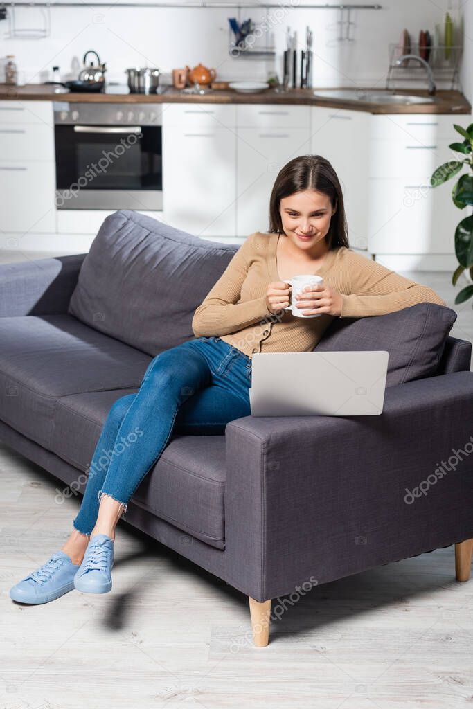 pleased freelancer holding cup of warm beverage while sitting on couch in kitchen near laptop