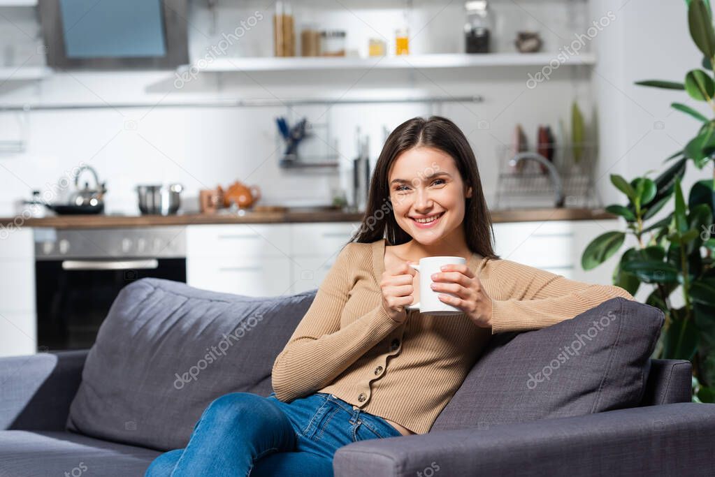 joyful woman looking at camera while sitting on sofa in kitchen with cup of warm tea