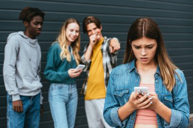 Seçici odak noktası, parmaklarını açık havada gösteren çoklu etnik kökenli gençlerin yanında akıllı telefon tutan üzgün genç. 