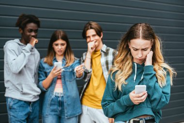 Açık havada gülen çoklu etnik gençlerin yanında akıllı telefon kullanan üzgün bir kızın seçici odağı. 