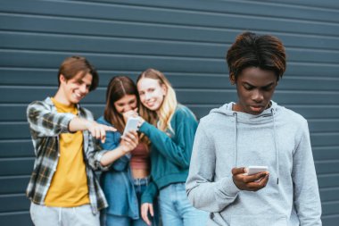 Açık havada gülen gençlerin yanında akıllı telefon kullanan Afrikalı Amerikalı çocuğun seçici odak noktası. 