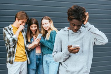 Açık havada cep telefonuyla gülümseyen gençlerin yanında akıllı telefon kullanan üzgün Afrikalı Amerikalı çocuğun seçici odak noktası. 