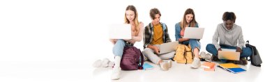 Panoramic shot of smiling multicultural teenagers using laptops near backpacks and books on white background clipart