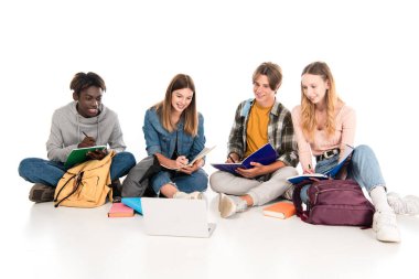 Gülümseyen çok kültürlü gençler beyaz arka planda dizüstü bilgisayarın yanında not defterlerine yazı yazıyorlar.