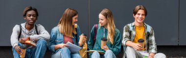 Panoramic shot of multiethnic teenagers with coffee to go and notebooks talking near building  clipart