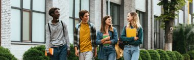 Panoramic shot of smiling multicultural teenage friends with notebooks walking on urban street  clipart