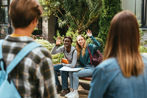 Selective focus of multiethnic teenagers with notebooks waving hands at friends on bench 