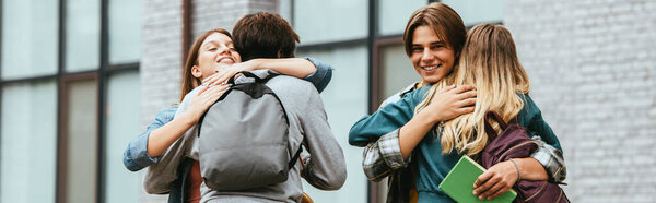 Panoramic orientation of smiling multicultural teenagers with backpacks embracing outdoors 