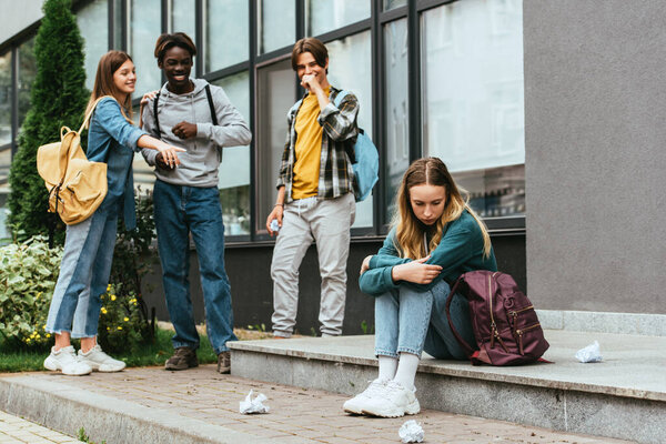 Selective focus of sad girl sitting near backpack, clumped paper and smiling multiethnic teenagers outdoors 