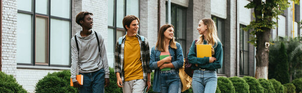 Панорамный снимок улыбающихся мультикультурных подростков с блокнотами, идущими по городской улице 