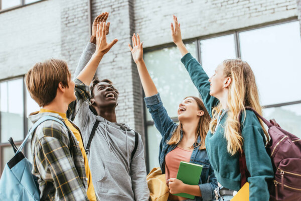 Селективный фокус позитивных мультикультурных подростков с рюкзаками и поднятыми руками на открытом воздухе 