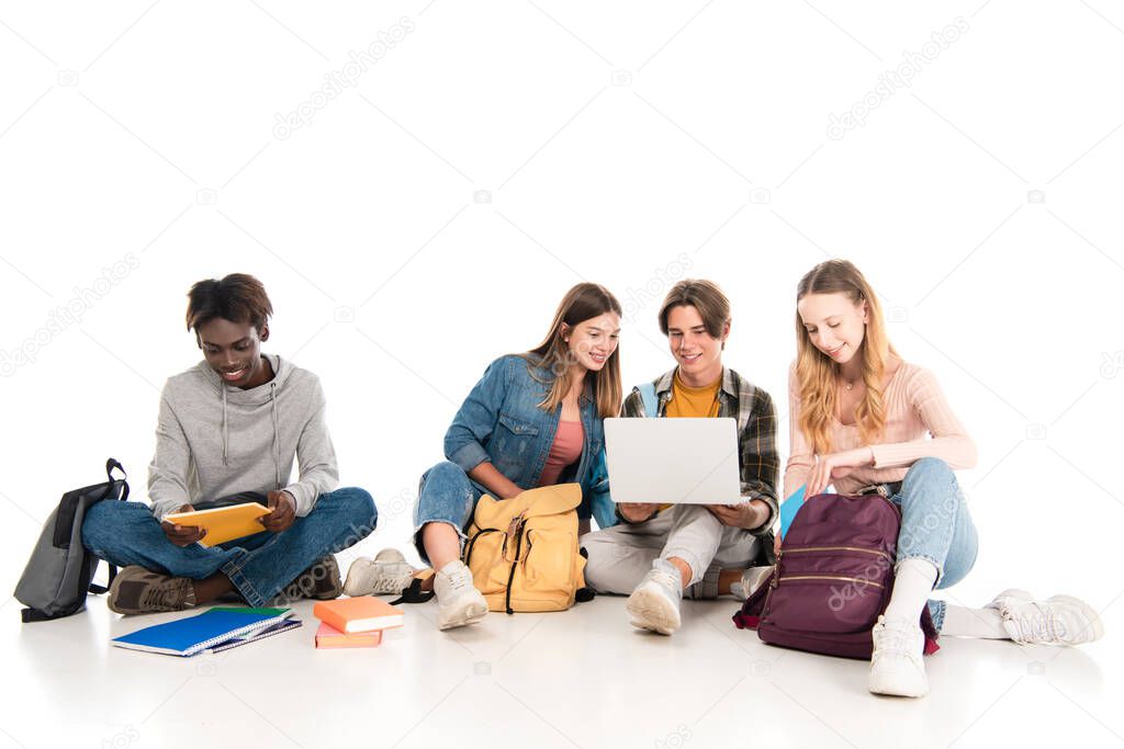 Smiling multicultural teenagers using laptop near books and backpacks on white background