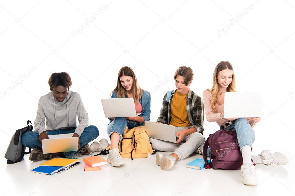 Positive multicultural teenagers using laptops near books and backpacks on white background