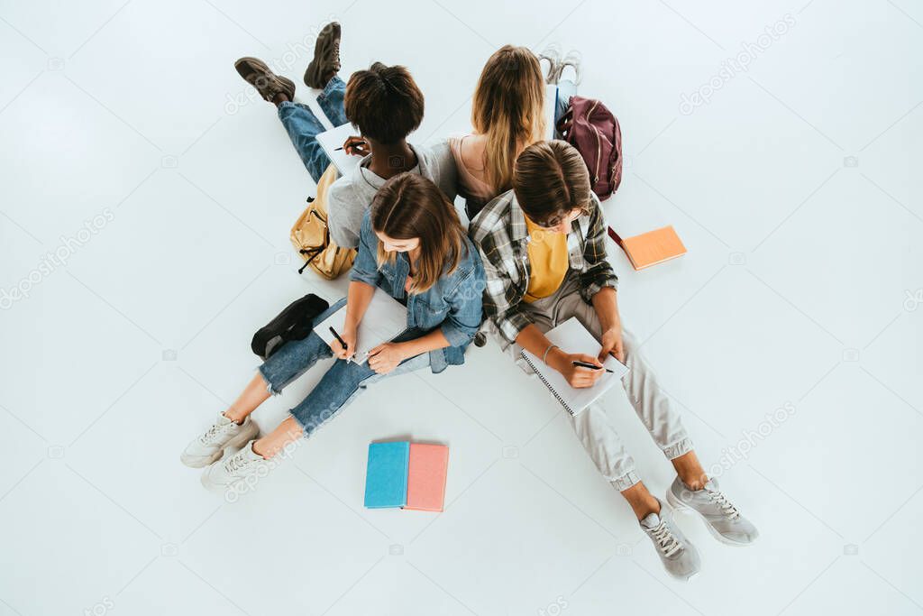 Overhead view of multiethnic teenagers writing on notebooks near backpacks  on grey background