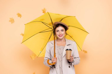 Trençkotlu ve bereli neşeli kadın şemsiyenin altına girmek için kahve tutuyor ve şeftalinin üzerine yapraklar döküyor.