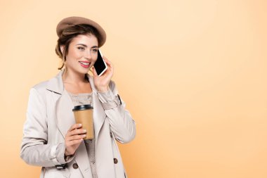 Şık bir sonbahar elbisesi içinde genç bir kadın şeftali ağacından izole edilmiş akıllı telefonuyla konuşurken elinde kahve tutuyor.