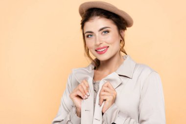 Bere takmış genç bir kadın şeftaliden izole edilmiş kameraya bakarken trençkotuna dokunuyor.