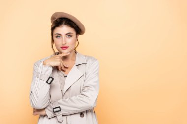 fashionable woman in trench coat and beret touching chin while looking at camera on peach clipart