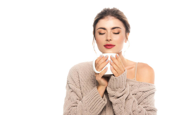 pleased woman in knitted sweater holding cup of warm beverage with closed eyes isolated on white
