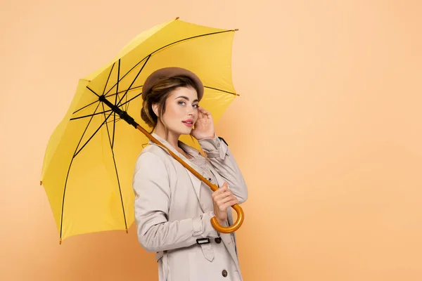 トレンチコートのファッショナブルな女性とベレー帽は桃の上に黄色の傘の下で顔 — ストック写真