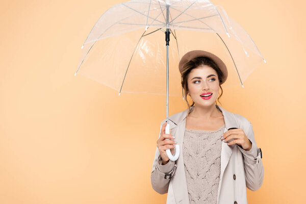 fashionable woman in trench coat and beret looking away under transparent umbrella on peach