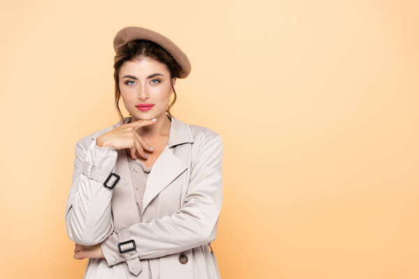 fashionable woman in trench coat and beret touching chin while looking at camera on peach