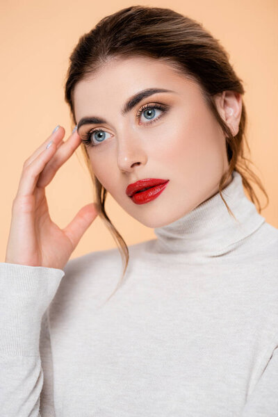 young woman in turtleneck touching face and looking away while posing isolated on peach 