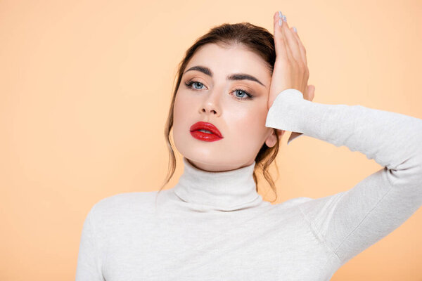 stylish woman in turtleneck looking away while touching face isolated on peach 