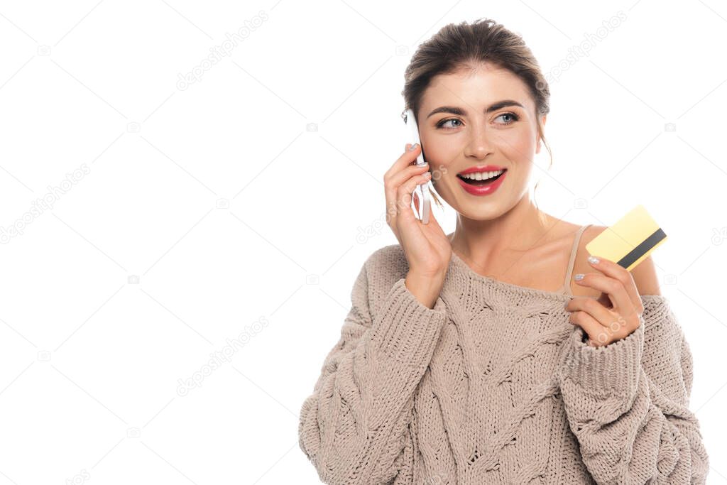 joyful woman in knitted sweater talking on smartphone while holding credit isolated on white